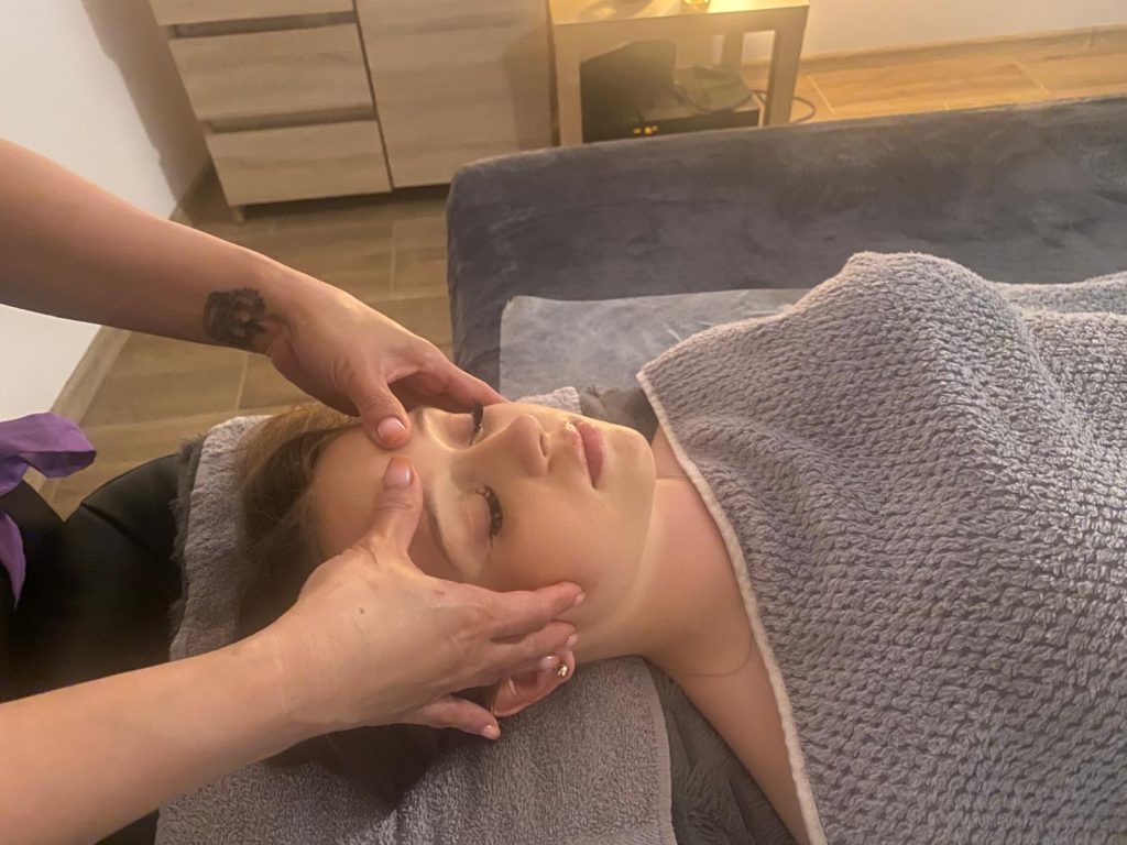 Kobieta leży na kozetce spa owinięta szarym ręcznikiem, podczas gdy kosmetyczka wykonuje jej masaż twarzy.