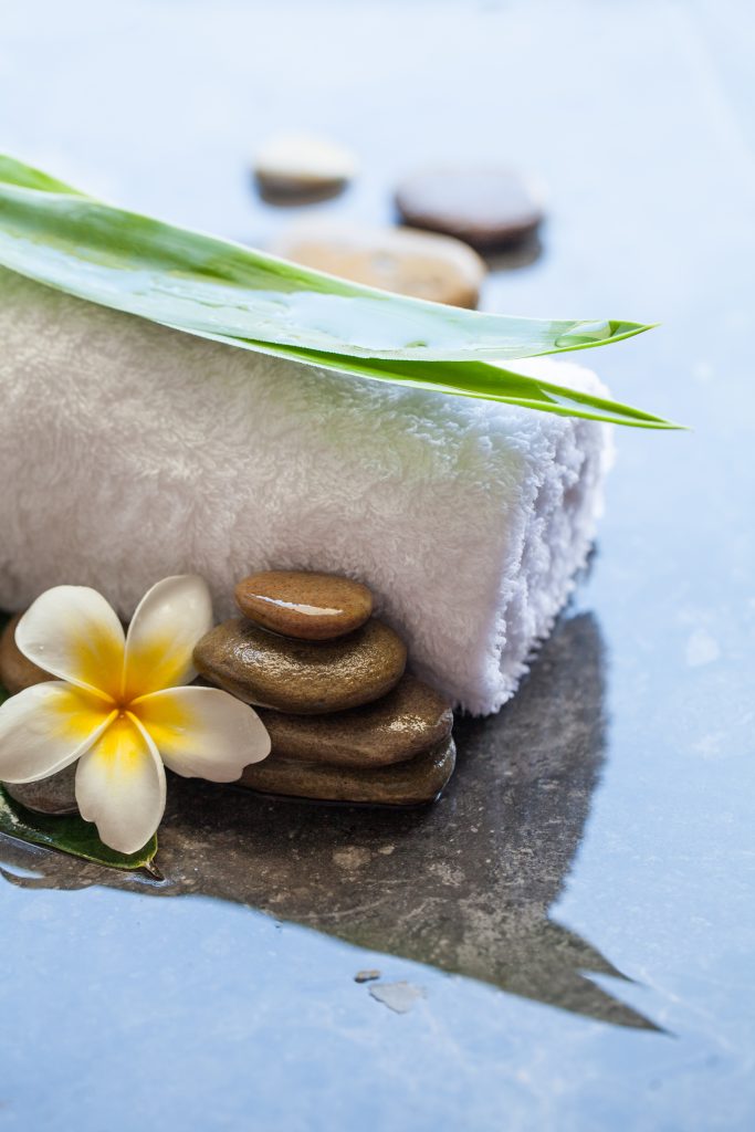 Zwinięty biały ręcznik, kwiat frangipani, zielone liście bambusa i ułożone kamienie spa na mokrej powierzchni.